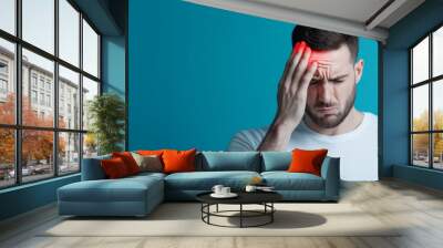 A young man in a white shirt experiencing a headache, holding his head in pain against a bright blue background. Wall mural