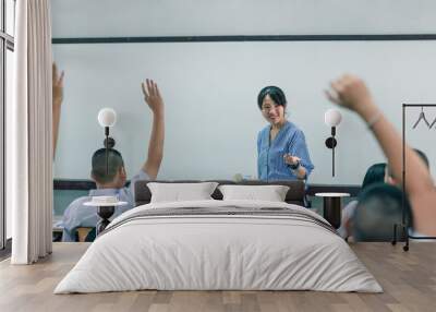 A smiling Asian female high school teacher teaches the white uniform students in the classroom by asking questions and then the students raise their hands for answers. Wall mural