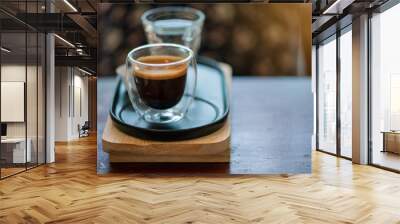 Hot espresso coffee in a clear glass, placed on a metal tray and wooden layer beside the coffee cup with a bear shaped cookie . All laid on dark brown wooden tables in retro coffee shop. Wall mural