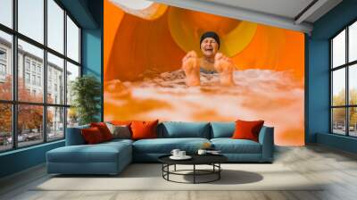 Fun swimming pool or aqua park activities. Laughing amused elderly caucasian female pensioner in black hair cap sliding on an orange water slide. Blurred feet in the foreground. High quality photo Wall mural