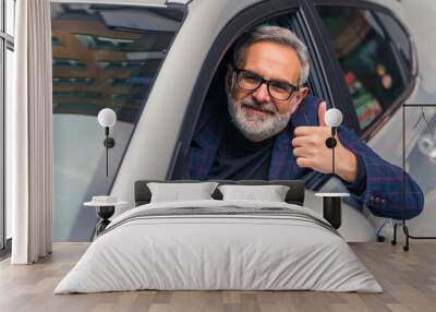 Handsome bearded mature man sitting in the drivers seat inside his newly bought car, leaning out the window, smiling joyfully, showing a thumb up. High quality photo Wall mural