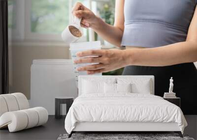 Young sporty woman pouring protein powder into a cup to make replacement food meal after workout Wall mural