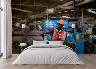 Close-up portrait of african-american man worker have unkempt beard in safety uniform, wearing hard hat, glasses, vest and glove are working on heavy machinery in industrial manufacturer factory. Wall mural