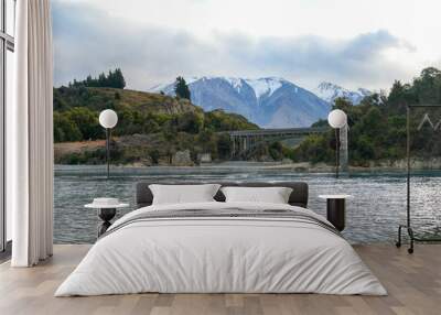 A scenic bridge crossing a river with snow-capped mountains in the background in New Zealand. The natural landscape includes trees and rocks surrounding the peaceful waters of the river. Wall mural