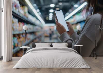 Female hand holding smartphone at supermarket and checking shopping list,blurred of store with trolley in department store,Mockup image of woman using mobile phone with blank white screen. Wall mural