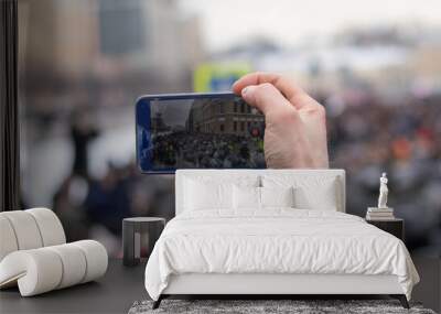 Hand with phone filming video. Police and protest demonstration on screen. Display with crowd of people, blurry background with copy space. Wall mural