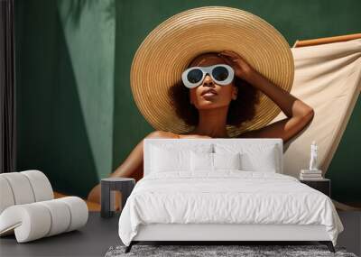 Black woman basks in sun, reclining on a sun lounger by pool in stylish hat and sunglasses Wall mural