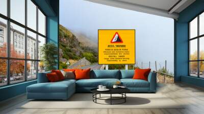 Yellow information sign warning of rockfall danger in Portuguese and English language. The usage of the road is under exclusive and only responsibility of the users. Misty landscape in the background Wall mural