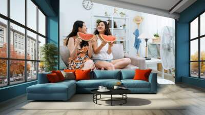 asian japanese female friends eating watermelon and relaxing on sofa in living room with electric fan blowing wind. young girls laughing holding slice of red fresh summer fruit with air cooler beside Wall mural