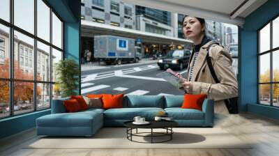 asian Japanese girl backpacker using map and navigation app on phone near a crosswalk in Kyoto japan. she looks into distance for traffic while waiting to cross the street Wall mural