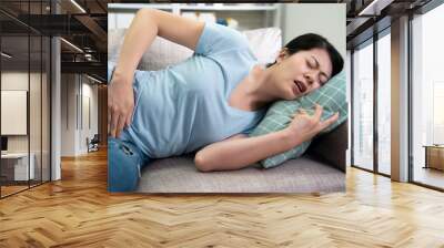 casual korean woman feel pain with menstruation lying on sofa hand catching pillow. asian mom at home on couch with stomachache. female worker resting in apartment with abdominal painful in day time. Wall mural