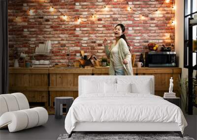 Cheerful young funny asian woman dancing and singing while having leisure time in kitchen at home. warm winter modern place apartment with red brick wall and hanging up light. elegant female relax. Wall mural