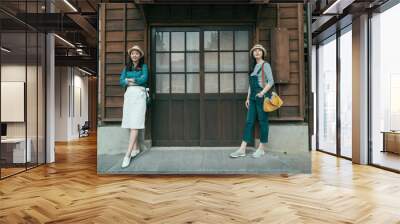 full length front view of two asian girl travelers standing by wooden door of traditional japanese ancient house. charming women tourists leaning on wood wall with copy space outdoor vintage style. Wall mural