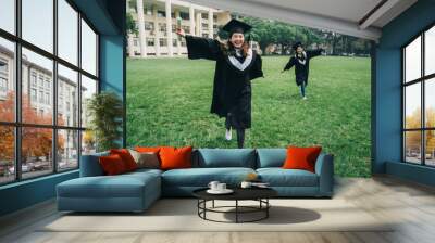 full length group of asian friends having fun running on green meadow in campus. two college girl students in mortar boards and bachelor gowns with diploma smiling carefree open arms relax sunshine Wall mural