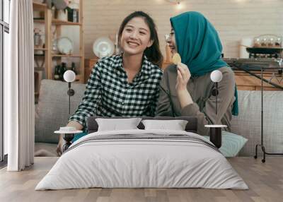 Happy multiethnic young women sitting on couch and watching funny movie on tv. asian girl roommates laughing with comedy playing on television and eating junk food in home kitchen on weekends. Wall mural