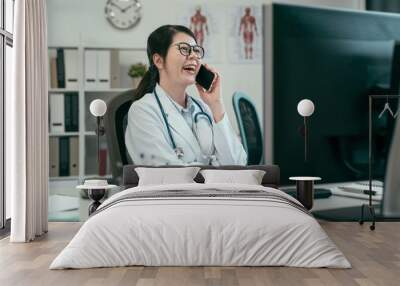 Smiling asian woman doctor talking on mobile phone at hospital. young female medical employee work in clinic office relax chatting on cellphone laughing gossip cheerful. happy lady having fun break Wall mural