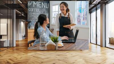 Smiling pretty girl serve coffee to female cafe visitor. happy young waitress bring ordered cappuccino and croissant to lady client working at laptop in coffee shop. woman freelancer chat with staff Wall mural