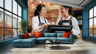 Team work and coworkers concept. two smiling female barista working together behind bar counter in coffeehouse. young girls coffee store staffs wear aprons using digital tablet talking and laughing. Wall mural