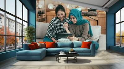 Two beautiful multiracial young women relaxing on sofa in living room. muslim lady with girl friend looking at digital tablet computer and laughing at something on screen. vintage style photo concept Wall mural