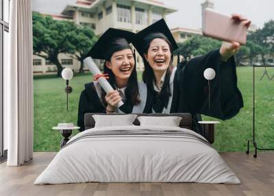 two cheerful asian college girl study abroad standing outdoor on lawn finish bachelor degree. Capturing happy moments. happy female student in graduation gowns making selfie smiling hold smart phone Wall mural