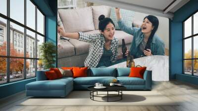 two female happy friends football fans watching soccer on tv and celebrating victory at home. Friendship sports and entertainment concept. asian women sitting on wooden floor drinking bottles of beer Wall mural