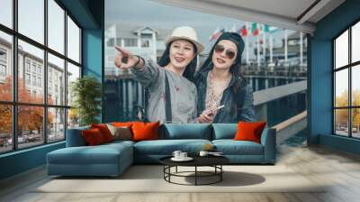 two happy asian women travel backpacker smiling with hats and cellphone cheerful point finger showing friend amazing ocean scenery while sightseeing in Stearns Wharf in Santa Barbara California usa. Wall mural