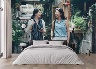 two happy girl friends chatting laughing relax walking by nature cafe shop surrounding by green plants trees. young women with smart phone searching way on internet online map app talking with sister Wall mural