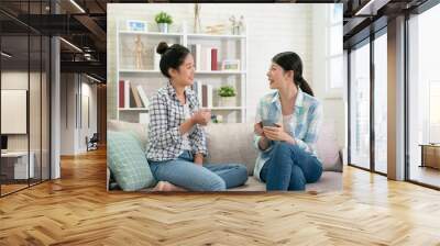 two happy young asian female friends with coffee cups conversing in living room at home. group of beautiful girls bestie sitting on couch relax in bright cozy apartment enjoy tea in weekends gossip Wall mural