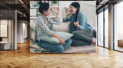 Two laughing asian female friends talking in living room at home sitting relaxing on couch. girls chatting about life and relations gossip and slumber party concept. women roommates having fun sofa Wall mural