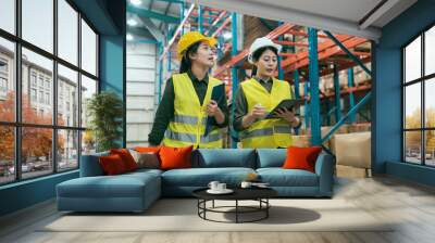 Two successful smiling businesswoman walking through big warehouse with helmets on their heads. woman manager holding digital tablet and colleague beside carry documents with clipboards in stockroom. Wall mural