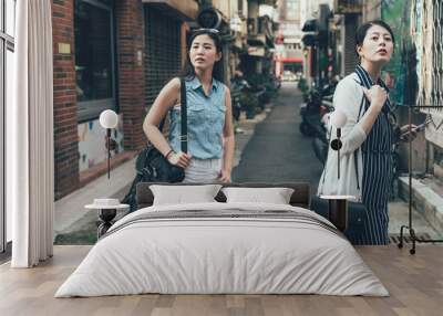 two young asian girl tourists consulting right way in city using smartphone gps in street searching locations. beautiful ladies travelers finding ways standing on road outdoor in urban look away Wall mural