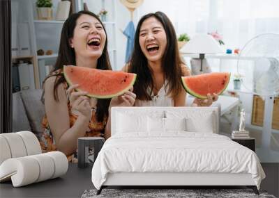 two young asian korean girls enjoying watermelon sitting on couch. Female friends eating fresh fruit slice and laughing together relax in living room with electric fan beside turning on breeze wind Wall mural