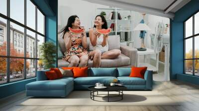 two young asian women with dress and shorts bite juicy watermelon. girl friends sitting on couch sofa at summer home indoor hot eating fresh fruit feeling cool in apartment with electric fan near. Wall mural