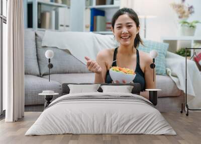 young asian elegant woman eating bowl of healthy salad after working out at home. happy girl face camera smiling while enjoy meal sitting on floor by sofa in home living room. hungry sporty girl. Wall mural