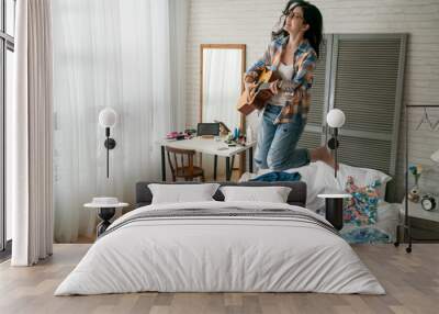 young funny girl playing acoustic guitar have fun dancing on bed at home. cheerful lady jumping in bedroom enjoy music singing songs in apartment with messy colorful clothes. happy female smiling Wall mural