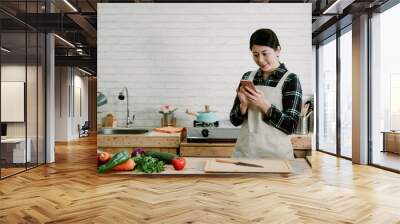 Young pretty asian chinese woman using smartphone standing at counter table full of fruits and vegetables in wooden kitchen interior. Counting calories with mobile phone app. Food health care concept Wall mural