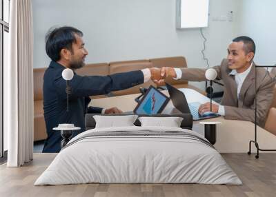 Two businessmen shaking hands in agreement while analyzing data on laptops, focusing on successful negotiation, collaboration, and business decision-making. Wall mural