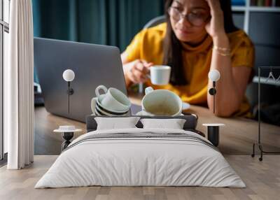 A piles of used coffee cups in front of an overtired Asian woman holding a cup of coffee sitting working frustrated exhausted looking at laptop screen. Caffeine addicted bad lifestyle concept.  Wall mural