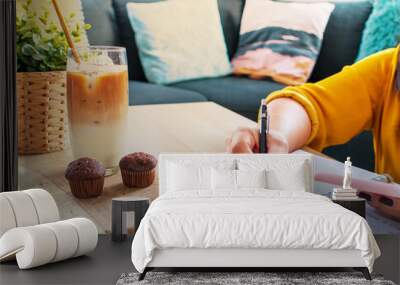 Cropped shot of young business female in yellow shirt writing short note and holding smartphone sitting with iced coffee and muffins comfortable working from home near the windows. Wall mural