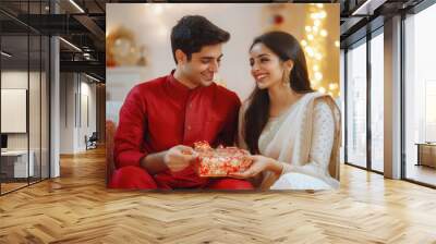 A handsome young Indian man in traditional wear giving a gift to his beautiful wife who wearing sari, sitting on a sofa. Both are smiling and happy Wall mural