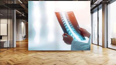 A doctor holds an X-ray of the spine up to the light, assessing it carefully in a bright clinical environment, highlighting attention to detail and patient care Wall mural