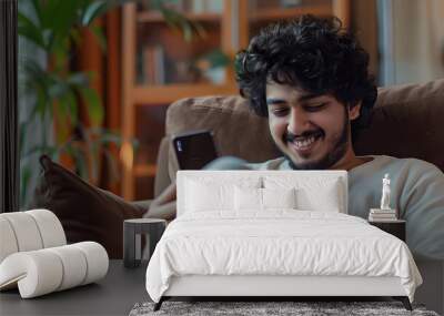 A happy curly young Indian man chats with girlfriend at home from a comfortable armchair, using a modern mobile phone to check social media and use a mobile app. There is copy space on the panorama. Wall mural