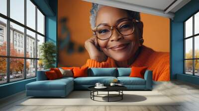 A joyful senior black woman wears stylish glasses and an orange sweater, with a warm smile that radiates happiness and confidence in a serene setting Wall mural