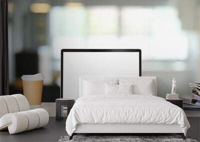 A white blank screen computer laptop is putting on a white table surrounded by a coffee cup and stack of books. Wall mural