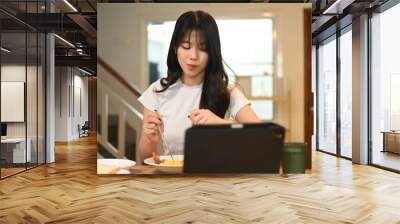 Attractive young woman enjoying her breakfast and using digital tablet at a kitchen table Wall mural