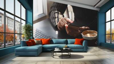 Cropped shot of businesswoman in formal striped suit holding smartphone in her hands in front a computer laptop and coffee cup while sitting at the modern working desk. Wall mural