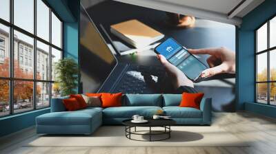 Cropped shot of executive woman holding smartphone in hands while doing online financial transaction by her smartphone in front computer tablet at the modern working desk. Wall mural