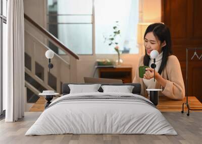 Happy young asian woman drinking hot coffee and using laptop on kitchen counter Wall mural