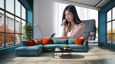 Young female entrepreneur having cell phone conversation and using laptop computer at office desk. Wall mural