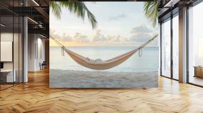 An inviting beachscape during the golden hour, showcasing a cozy setup with a hammock tied between two palm trees and a backdrop of a serene ocean sunset. The soft colors of the sky blend seamlessly Wall mural